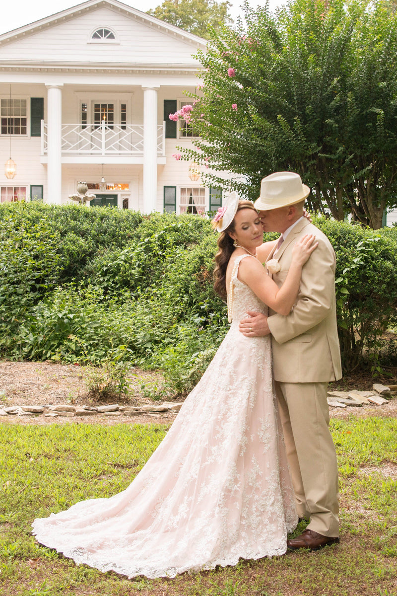 A bride in a Desiree Spice blush lace wedding gown and a man in a beige suit and hat embrace on a green lawn, with a large white house and manicured hedges in the background.