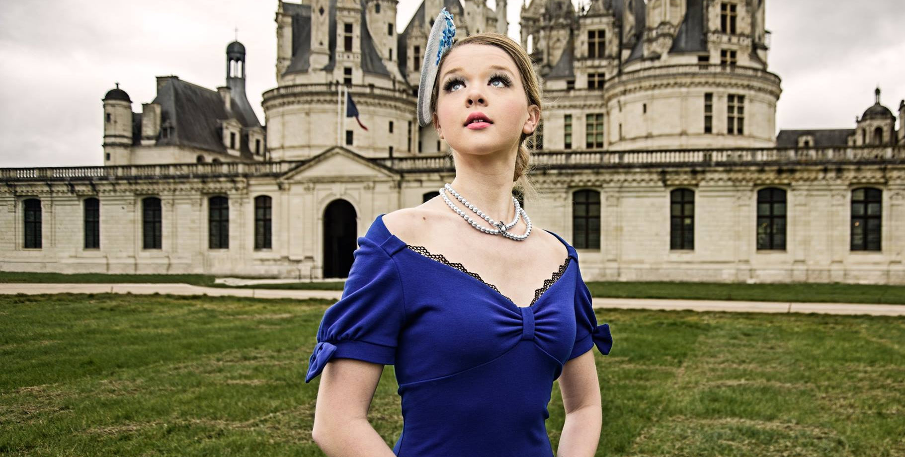 A woman wearing a Desiree Spice royal blue off-the-shoulder dress and a matching blue saucer fascinator hat stands on a grassy lawn in front of a large historic castle under an overcast sky.