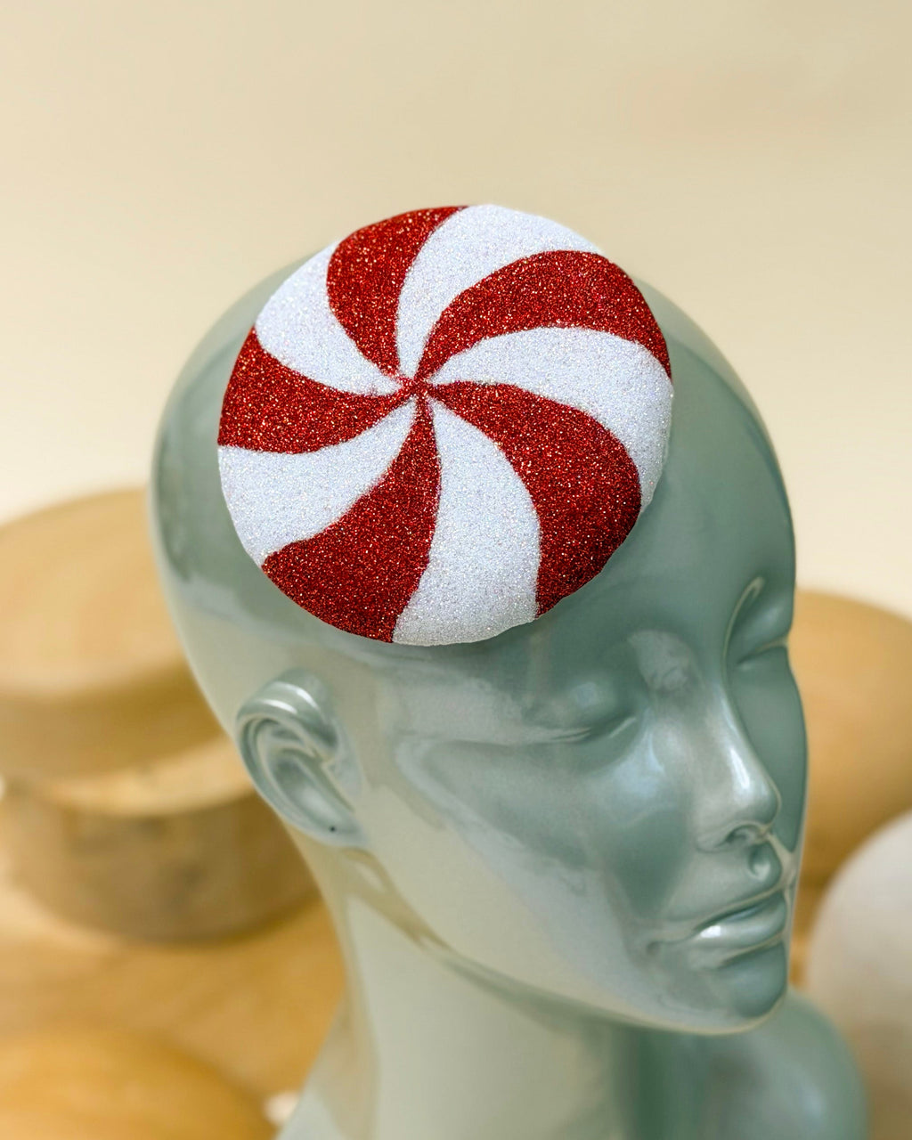 A red and white Christmas peppermint swirl glitter cocktail hat by Desiree Spice displayed on a light blue mannequin head, with wooden millinery blocks in the blurred background.
