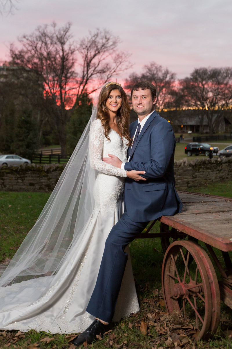 A bride in a couture Desiree Spice white lace wedding gown with a long sheer veil and a groom in a navy blue suit sitting on a rustic wooden cart outdoors at sunset, with trees and parked cars in the background.