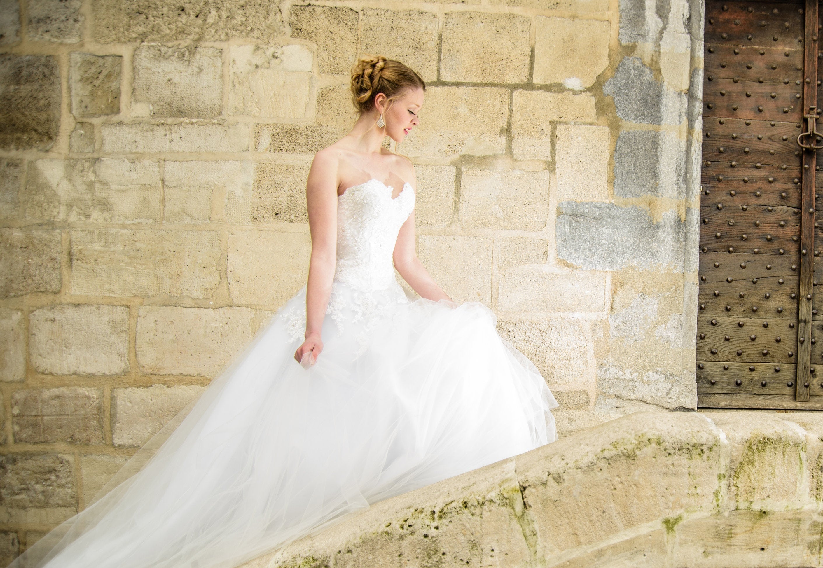 A bride in a strapless white Desiree Spice wedding gown with a full tulle skirt walks up a stone ledged staircase beside a stone wall and a dark wooden door.