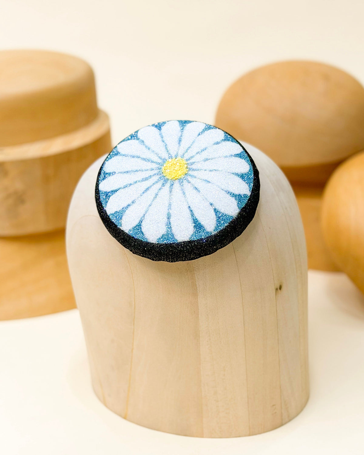 Glitter pillbox fascinator hat designed to resemble a daisy in glitter of black, blue, white, and yellow by Desiree Spice. Displayed on top of a wooden millinery block.