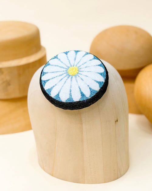 Glitter pillbox fascinator hat designed to resemble a daisy in glitter of black, blue, white, and yellow by Desiree Spice. Displayed on top of a wooden millinery block.