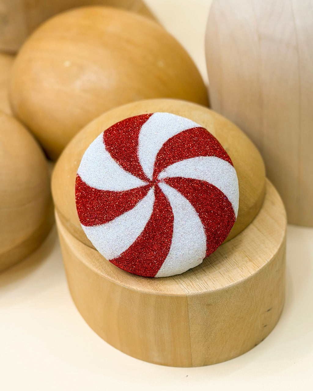 A Christmas peppermint fascinator cocktail hat by Desiree Spice featuring a red and white glittery peppermint swirl design, displayed on a small wooden millinery stand among other wooden decorative objects.