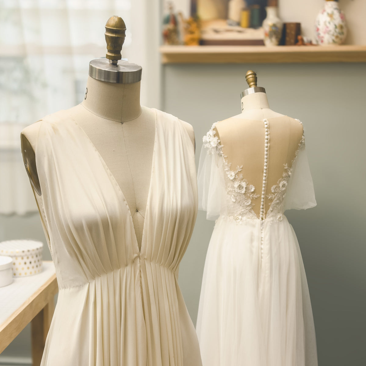 Two cream-colored wedding dresses displayed on mannequins in a softly lit boutique. The front view shows a sleeveless V-neck gown with pleated fabric, and the back view shows a white tulle dress with floral lace and pearl embellishments.