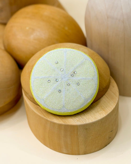 A glitter fascinator pillbox hat designed to look like a lemon slice by Desiree Spice, featuring a yellow center with white, a bright yellow outer edge, and Austrian crystals, displayed on a wooden millinery block with other wooden props.