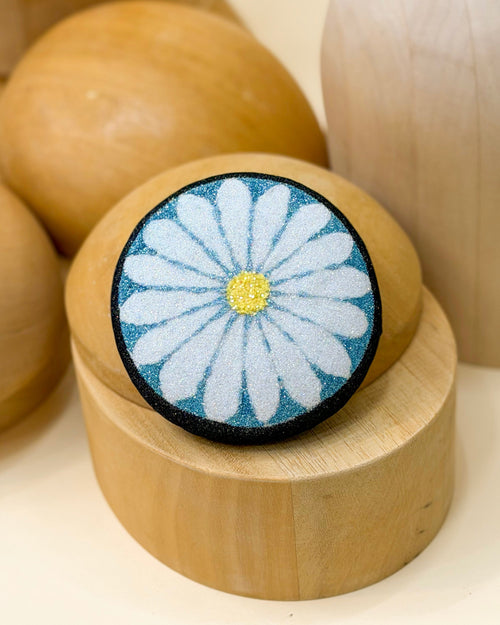Glitter pillbox fascinator hat designed to resemble a daisy in glitter of black, blue, white, and yellow by Desiree Spice. Displayed on top of a wooden millinery block.