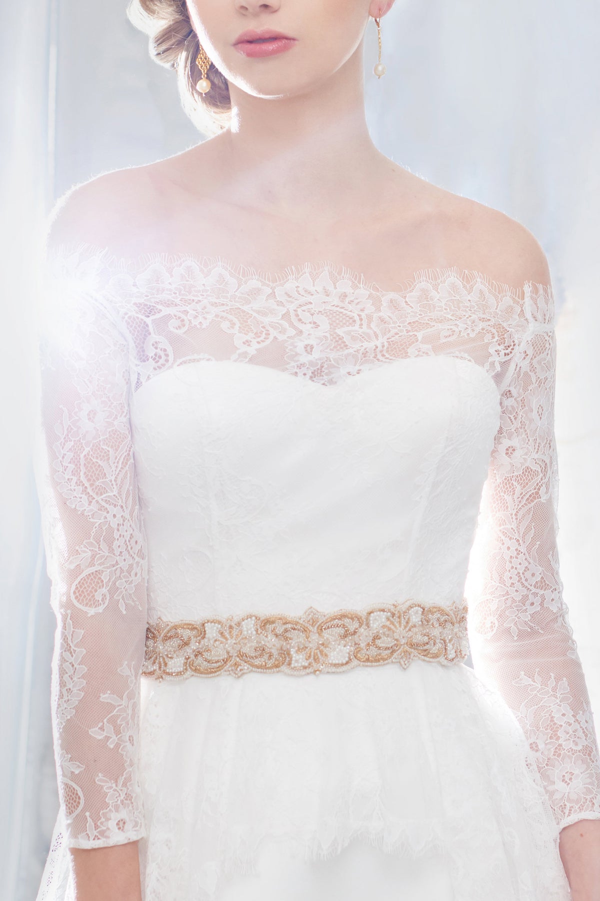 A close-up of a white Desiree Spice bridal gown with sheer lace long sleeves, an off-the-shoulder lace neckline, and an ornate gold embroidered waistband sash, shown on a model against a bright white background.