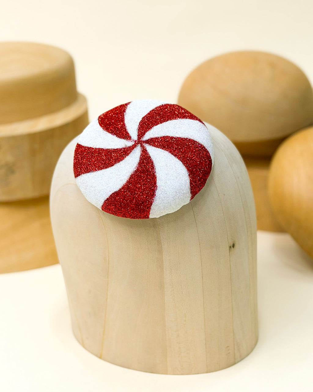 A Christmas fascinator hat with a red and white glitter peppermint swirl design by Desiree spice, displayed on top of a light wooden millinery blocks