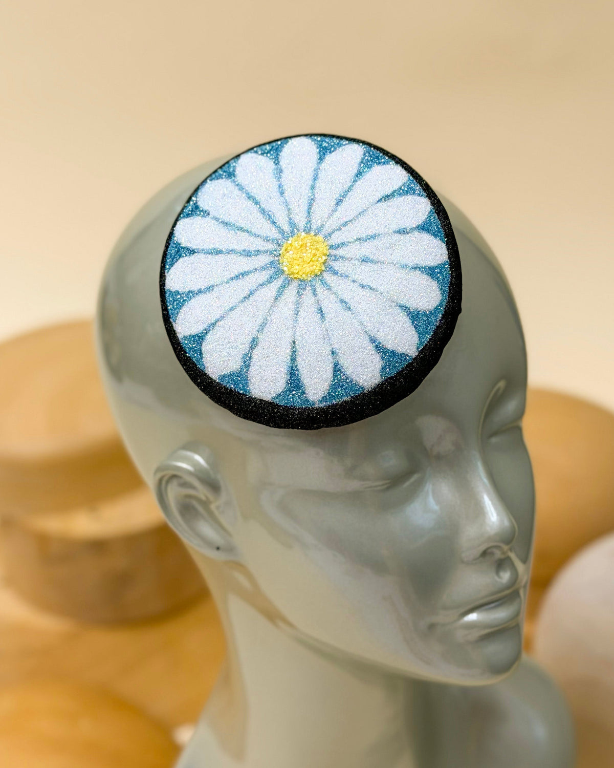 Glitter pillbox fascinator hat designed to resemble a daisy in glitter of black, blue, white, and yellow by Desiree Spice. Displayed on top of a wooden millinery block.