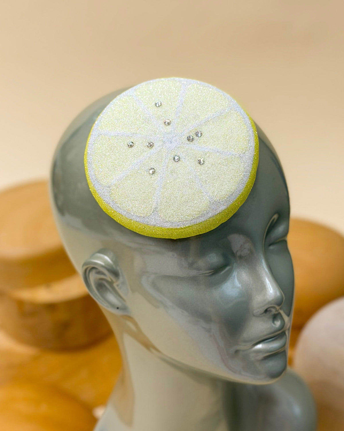 A yellow glitter fascinator hat resembling a lemon slice with Austrian crystals by Desiree spice, displayed on a gray mannequin head. The background features blurred wooden millinery hat blocks against a warm beige backdrop.