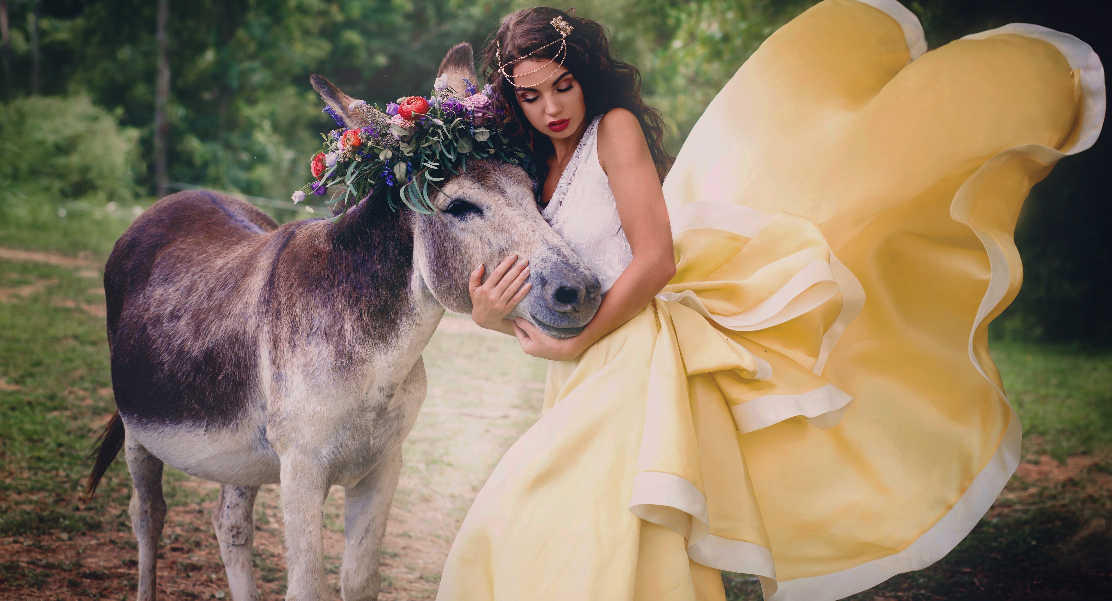 A woman in a flowing pale yellow bridal Desiree Spice couture gown embraces a donkey outdoors, with a floral crown on the donkey and a softly blurred forest background.